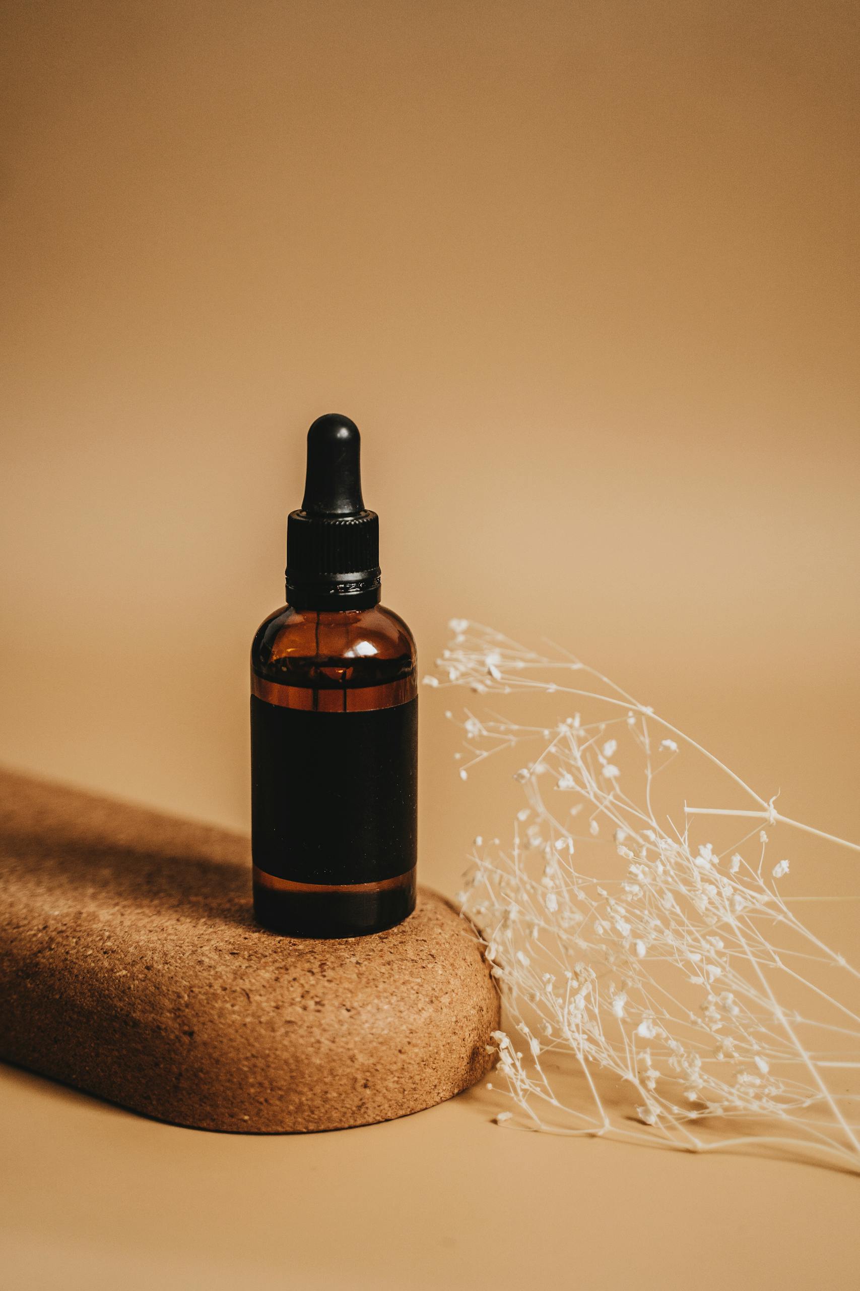 A stylish amber glass bottle with dropper sits on a natural cork surface with delicate dried flowers.